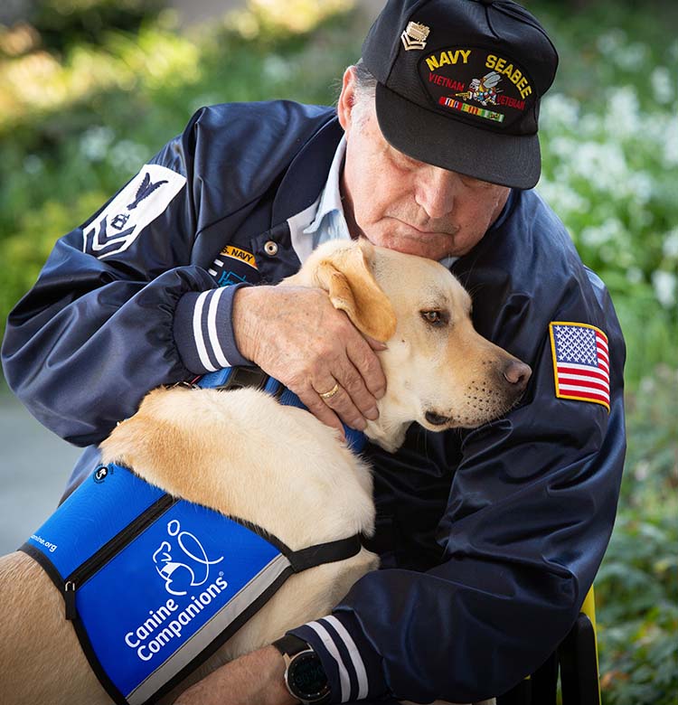 Man wearing a veterans hat and jacket hugging a white Labrador