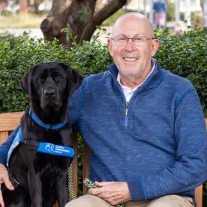 a smiling man sits next to a black lab service dog on a bench