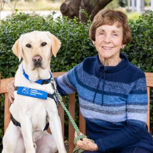 a smiling woman on a bench sitting next to a yellow lab service dog