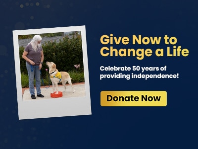 A woman with long gray hair is standing outdoors with a yellow dog, wearing a harness, next to a red bowl. Text on the image encourages donations to support independence.