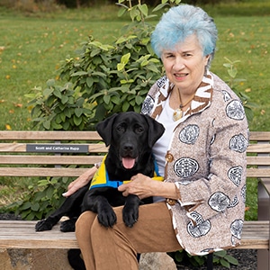 A smiling woman sitting next to a black lab puppy in a yellow puppy vest