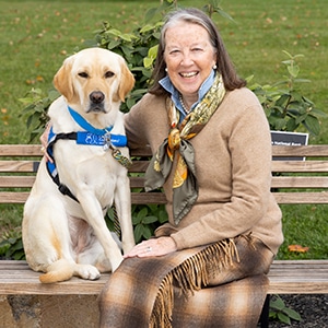 A smiling woman sitting next to a yellow lab service dog in a blue canine companions vest