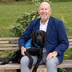 A smiling man sitting next to a black lab service dog in a blue canine companions vest