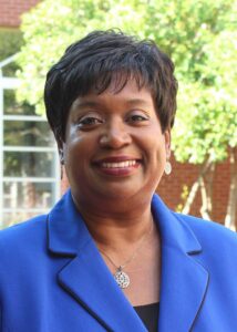 Dr. Teresa Johnson Ramey: Smiling woman wearing a blue blazer and a pendant necklace, with short hair standing outdoors in front of trees and a building.