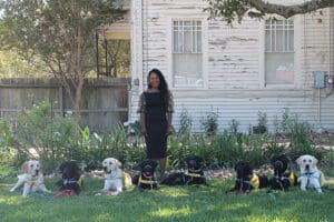 Tanisha Hall standing in front of a house with seven Labrador retrievers wearing yellow vests sitting on the grass.