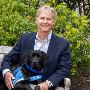 A man sitting on a bench outdoors with a black lab in a blue service vest laying across his lap