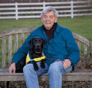 A smiling man on a bench with a black lab puppy in a yellow puppy vest in his lap