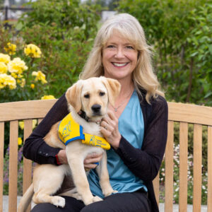 A smiling woman with a yellow lab puppy in a yellow puppy vest sitting on a bench outside among some flowers