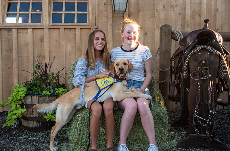 two girls sitting on hay bales next to saddle with yellow dog wearing a Canine Companions yellow puppy vest accross their laps