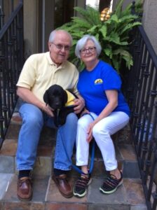 Older man and woman sit on an out door staircase, holding a black lab puppy in yellow puppy vest
