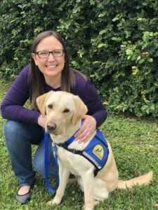Woman crouches next to a yellow lab service dog in blue vest
