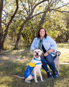 Woman sits next to a yellow lab service dog in blue vest