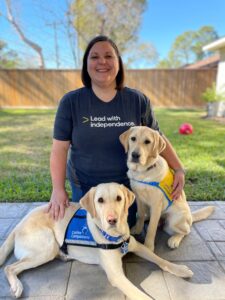 A woman kneeling with a yellow service dog and a yellow puppy