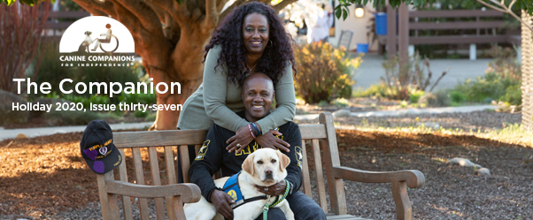 person sitting on a bench with a Canine Companions service dog across their lap and another person standing behind them.
