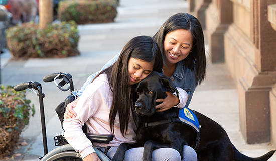 girl sitting in wheelchair with an adult next to her and a Canine Companions service dog over her lap