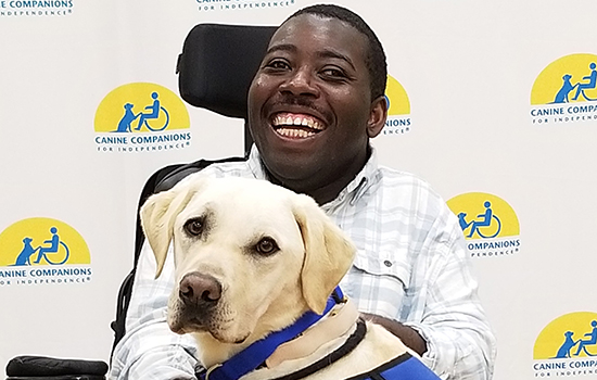 A young Black man using a power wheelchair smiles with a yellow Labrador in a blue vest resting its upper body on the man’s legs.