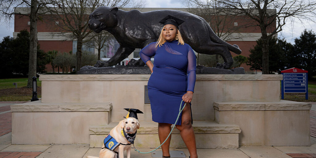 A woman in a blue dress and graduation hat stands next to a yellow lab in a blue service vest also wearing a graduation hat