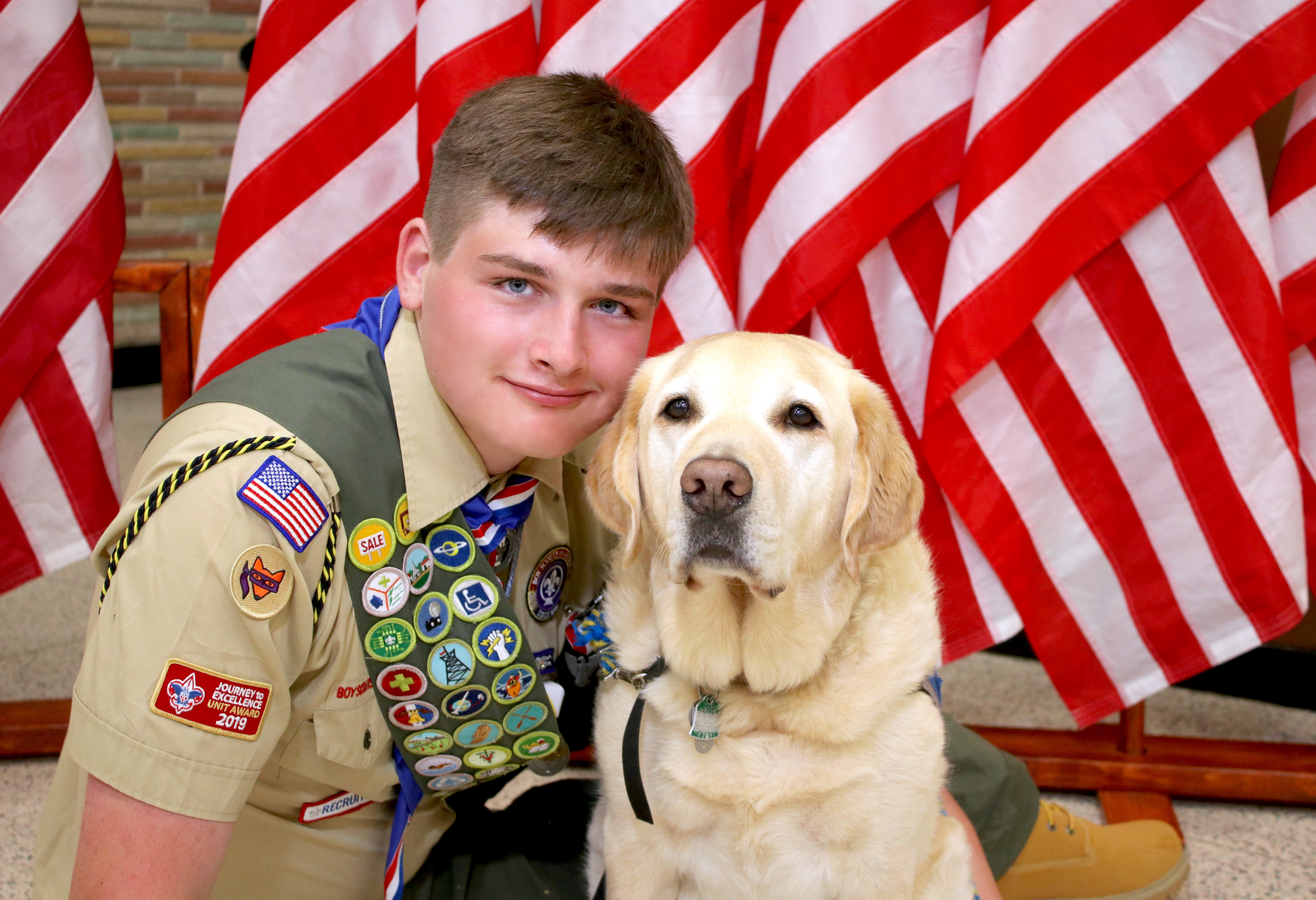 A young man in a Boy Scout uniform sits next to his yellow Labrador service dog with American flags in the background.