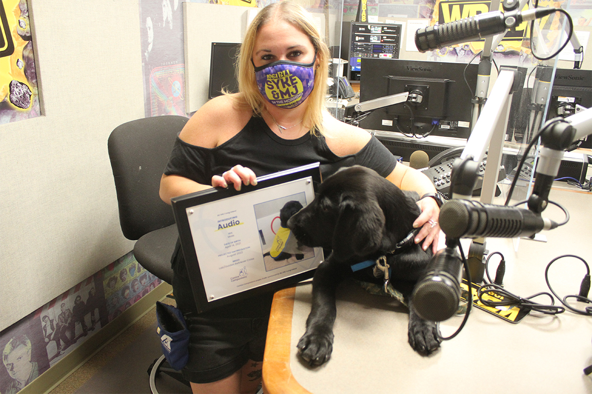 A woman holds a young black Labrador in a service dog vest in a radio broadcast room