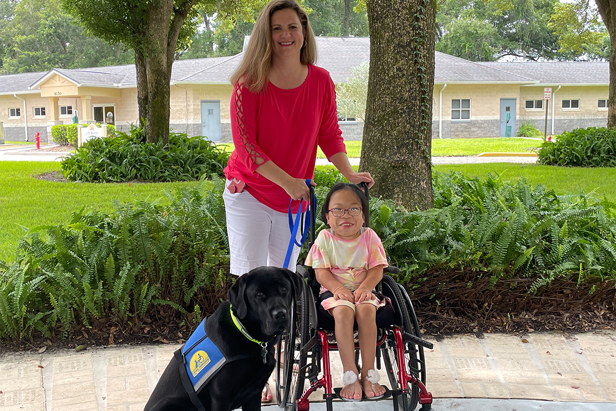 A young Asian girl in a wheelchair smile with her parent and a black Labrador service dog
