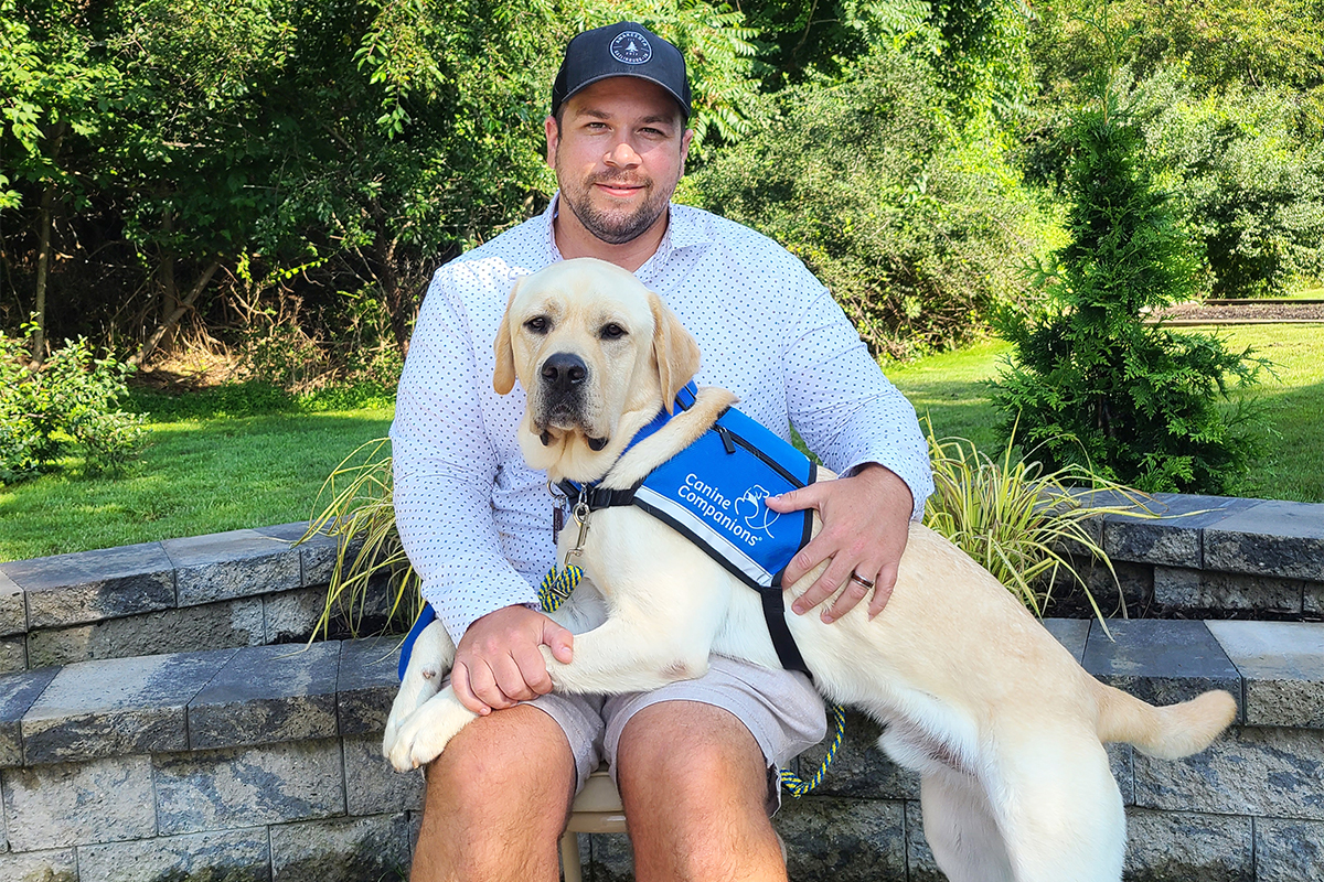 A man sits with a large yellow Labrador service dog in a blue vest on his lap.