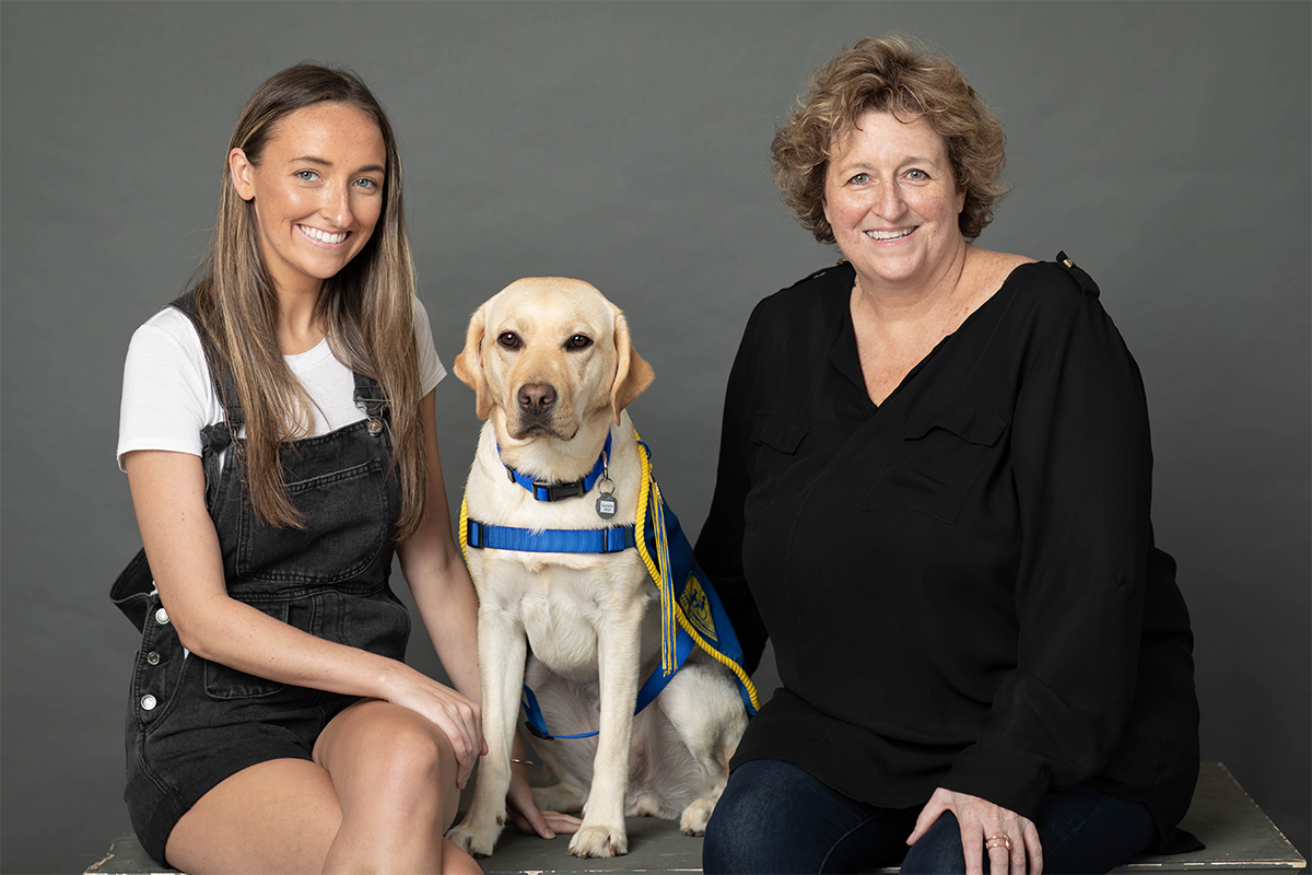 A young white woman sits in a black dress next to a yellow Labrador service dog and another woman in a black outfit