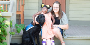 Woman sitting on steps holding a girl with glasses with her arm around a black Labrador’s back who is wearing a blue vest with Canine Companions text.