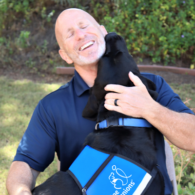 Man smiling and holding a black Labrador wearing a blue vest