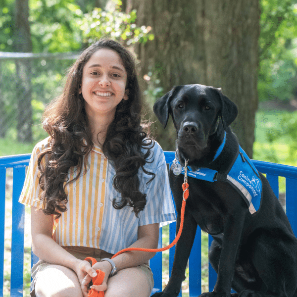 A young woman with long, curly hair wearing a striped shirt and shorts smiles while sitting on a blue bench next to a black dog wearing a blue harness, in a green outdoor setting.