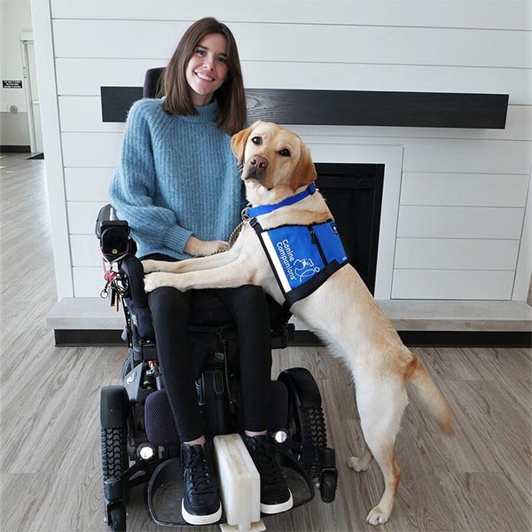 A young woman in a wheelchair smiles as her yellow lab in a blue service dog vest lays across her lap