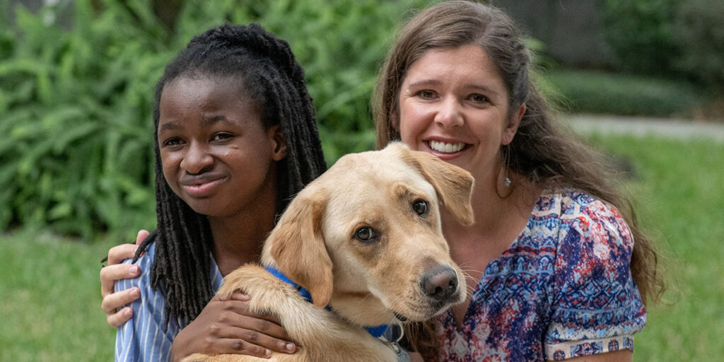 Two girls smiling with a Labrador wearing a blue vest