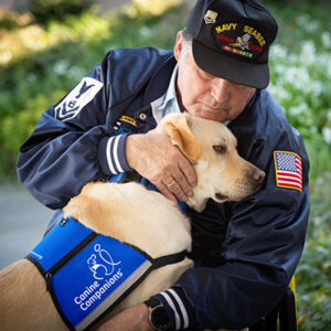 Man wearing a veterans hat and jacket hugging a white Labrador