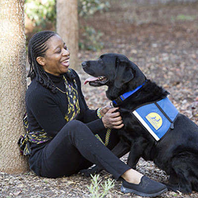 An African American woman sits against a tree petting a lab service dog in a blue vest