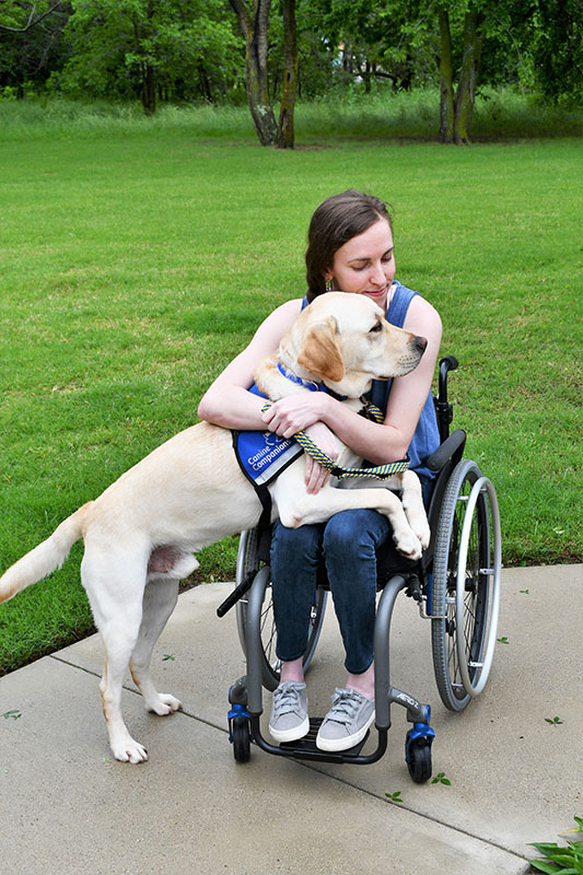 woman in a wheelchair hugging a yellow lab in blue service vest who is laying across her lap