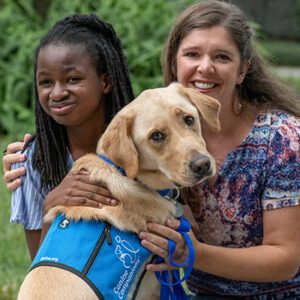 Mother and daughter smiling with a Labrador wearing a blue vest