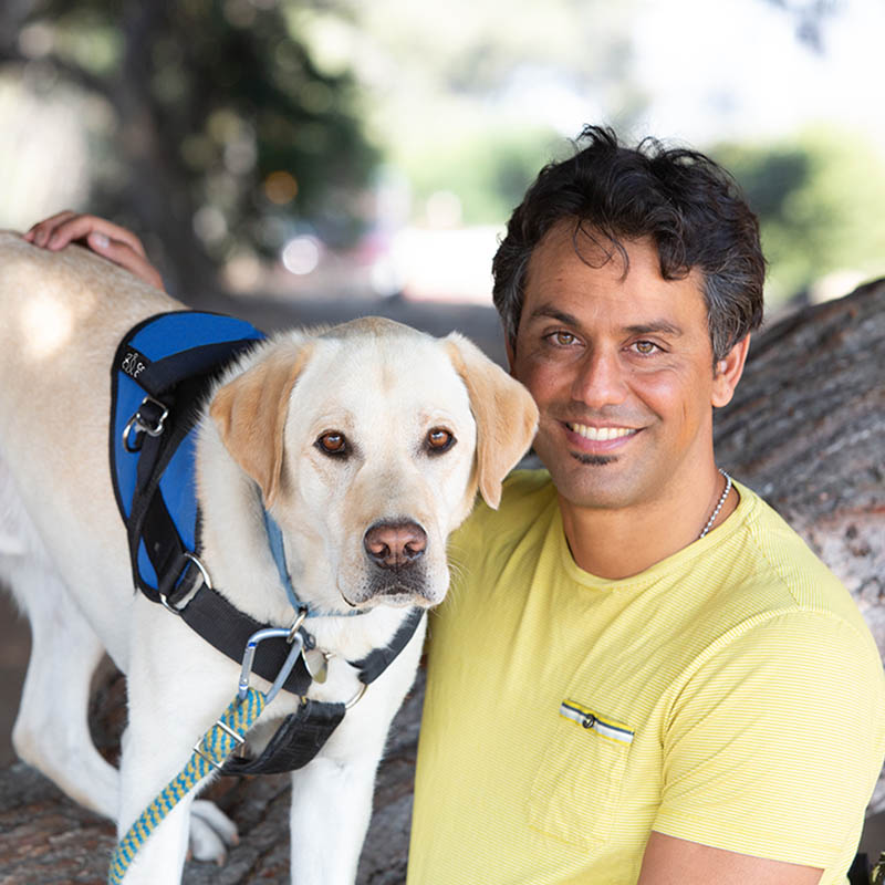 Man smiling with his arm around yellow lab in blue service vest, both leaning on a tree