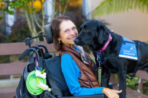 A woman in a wheelchair smiles with her black service dog next to her