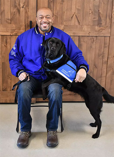 smiling african american man sits in a chair with his black lab service dog in blue vest