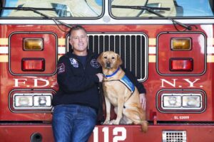 Man and yellow lab in a blue service vest standing in front of a FDNY fire engine