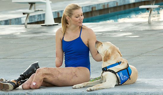 Woman sitting by a lap pool in a bathing suit with a yellow lab in a blue vest laying nearby