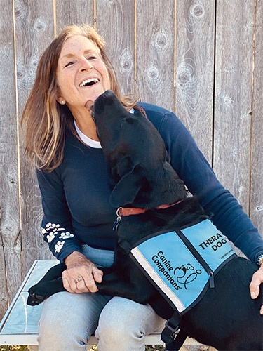 Woman sitting on a bench with a black lab in a teal Therapy Dog vest in her lap licking her face