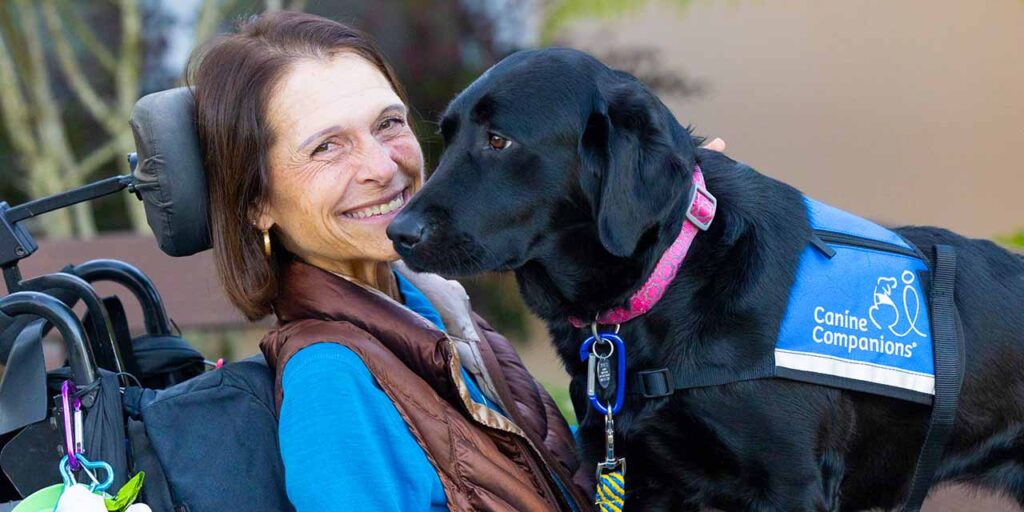 a woman on a wheelchair smiling, with a black Labrador standing on her lap wearing a blue vest with Canine Companions text