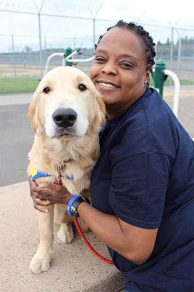 Woman in a prison yard hugs a golden retriever puppy in a yellow puppy vest