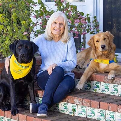 Woman sitting on brick steps next to a black Labrador and a brown Golden Retriever, both wearing yellow and blue gear