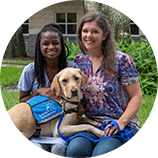 young girl with her mother sits with their yellow lab service dog wearing a blue vest