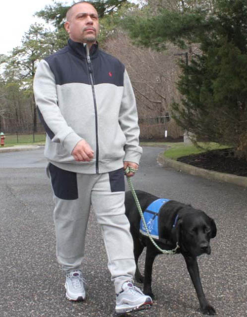 Man in a gray track suit walks along a road with his black lab in blue service vest