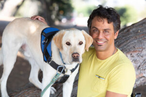 Smiling man in a yellow tshirt sits beneath a tree with his arm around his yellow lab service dog wearing a blue service vest