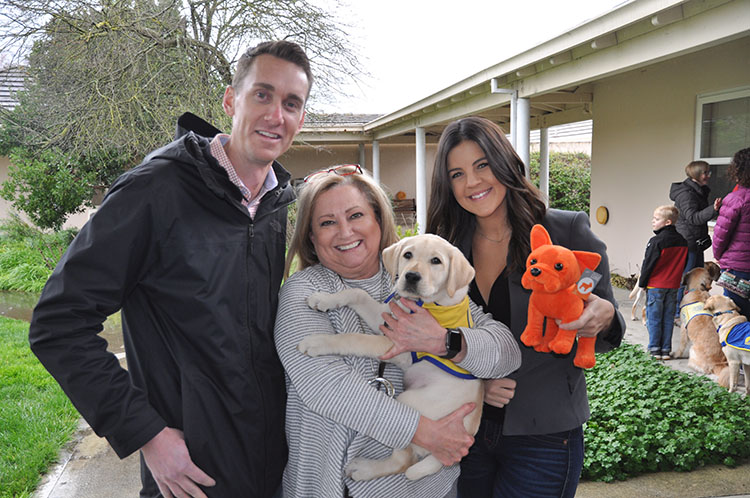 three people hold a puppy in a yellow puppy vest and a small orange nexthome mascot stuffed dog