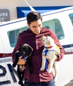 A young man holds two puppies in yellow puppy vests and stands in front of his small airplane