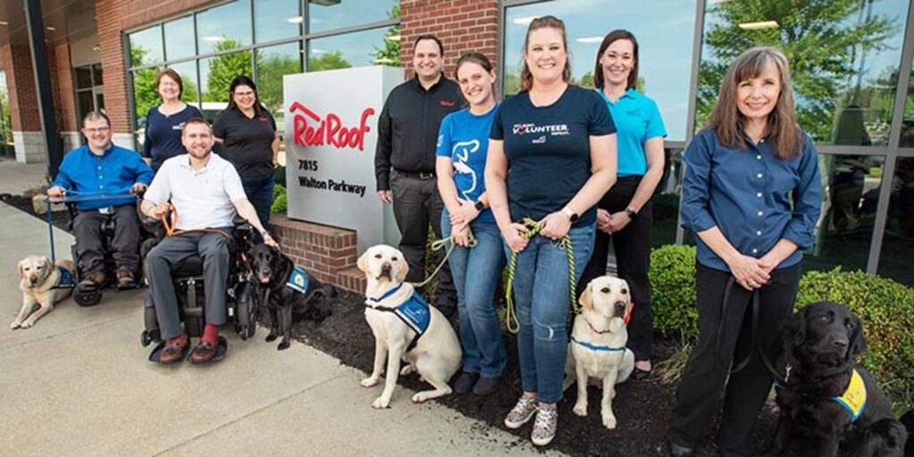 a group of graduates stand in front of the red roof corporate office with their service dogs in blue vests
